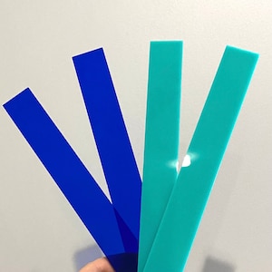 Puede incluir: Tres tiras largas y delgadas de plástico, una azul y dos turquesa, se sostienen en una mano. Las tiras son translúcidas y tienen un ligero brillo.