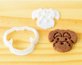 May include: Two white plastic cookie cutters shaped like a dog's head, and a brown dog-shaped cookie.
