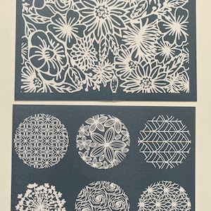 May include: Two sheets of white vinyl stencils with floral and geometric designs. The top sheet features a large floral pattern with various flower shapes and sizes. The bottom sheet has six circular stencils with different patterns, including a floral design, a geometric pattern, and a design resembling a starburst.
