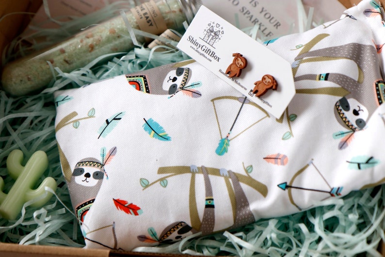 May include: A white fabric pouch with a sloth pattern. The pouch is filled with green shredded paper. The pouch has a brown sloth with a headdress and feathers. The pouch also has a brown sloth with a bow and arrow. The pouch is in a box with a green shredded paper background. The pouch is a gift box.