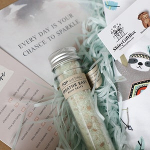 May include: A self-care gift box with a white and blue sloth design. The box includes a small glass bottle of bath salts labeled "Breathe Easy" and a card with a list of self-care activities. The card reads "Time for Self-Care" and includes activities like "Daily mood tracker", "Afternoon nap", "Time with family", and "Scented candles".