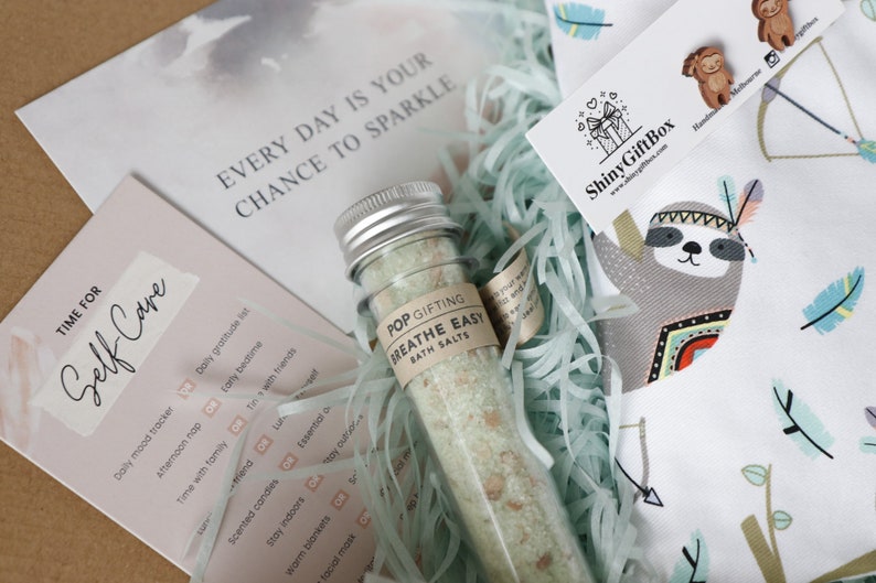 May include: A self-care gift box with a white and blue sloth design. The box includes a small glass bottle of bath salts labeled "Breathe Easy" and a card with a list of self-care activities. The card reads "Time for Self-Care" and includes activities like "Daily mood tracker", "Afternoon nap", "Time with family", and "Scented candles".