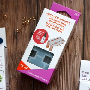 May include: A pink and white box with a clear plastic window showing a silicone mold for making jewelry. The mold is shaped like a table with four legs. The box also includes a small bottle of Liquid Sculpey, a brand of polymer clay. The text on the box reads "Oven-Safe Silicone Mold" and "3 pc|pz 3D Jewelry".