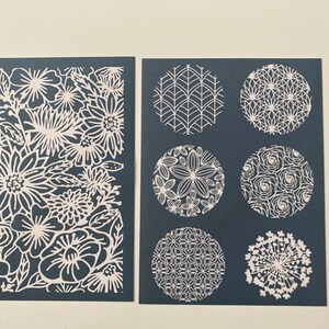 May include: Two cards with white paper cut designs on a dark blue background. One card features a floral pattern and the other card features six different circular patterns.