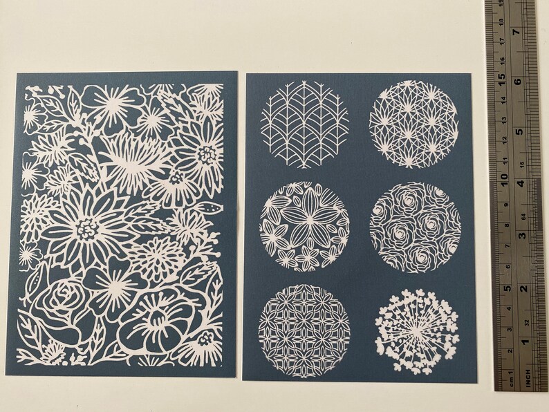 May include: Two cards with white paper cut designs on a dark blue background. One card features a floral pattern and the other card features six different circular patterns.