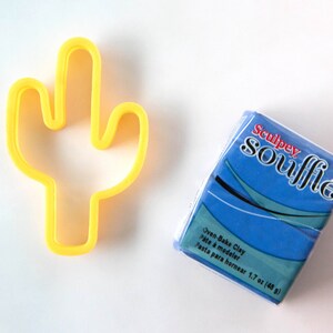 Puede incluir: Cortador de galletas de cactus de pl&aacute;stico amarillo y un paquete azul de arcilla para hornear Sculpey Souffle. El paquete dice "Oven-Bake Clay, P&acirc;te &agrave; modeler, Pasta para hornear 1.7 oz (48 g)"