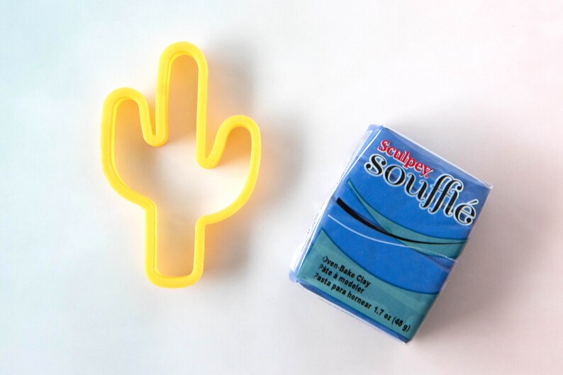 Puede incluir: Cortador de galletas de cactus de pl&aacute;stico amarillo y un paquete azul de arcilla para hornear Sculpey Souffle. El paquete dice "Oven-Bake Clay, P&acirc;te &agrave; modeler, Pasta para hornear 1.7 oz (48 g)"