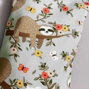 May include: A gray fabric eye pillow with a repeating pattern of sloths hanging from branches with flowers and butterflies. The sloths are brown with white faces and black eyes.