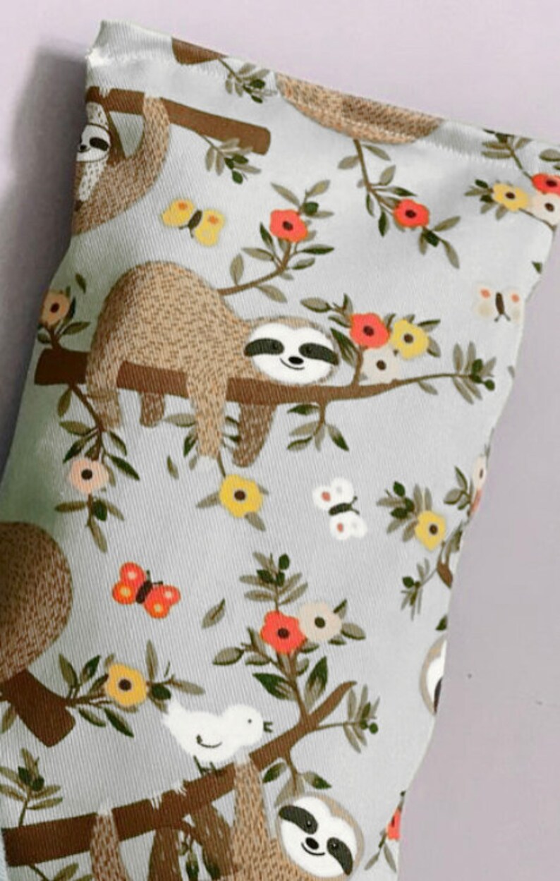 May include: A gray fabric eye pillow with a repeating pattern of sloths hanging from branches with flowers and butterflies. The sloths are brown with white faces and black eyes.