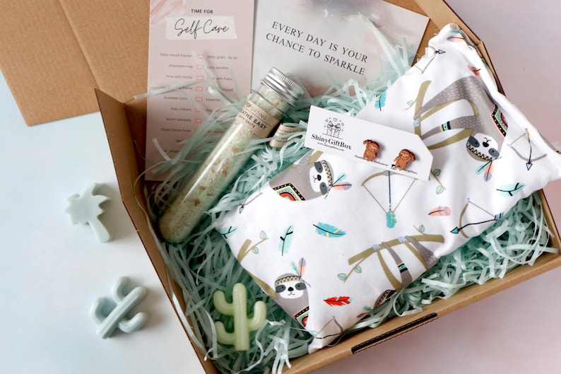 May include: A brown cardboard box filled with a self-care gift set. The box contains bath salts in a glass tube, a white fabric pouch with a sloth design, a pair of wooden sloth earrings, and two small green soap sculptures shaped like a palm tree and a cactus. The box is filled with green shredded paper. The text on the card reads "Time for Self Care" and "Every day is your chance to sparkle".