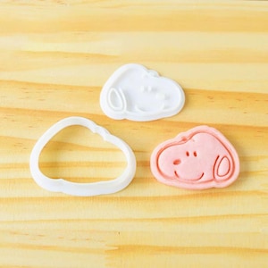 May include: Three Snoopy-shaped cookie cutters. One is a white outline, another is a white Snoopy face, and the third is a pink Snoopy face with details. The cutters are on a light wood surface.