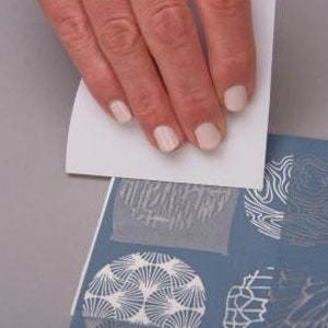 May include: A hand is pressing a white stencil onto a blue surface with white patterned designs. The stencil has a geometric pattern.