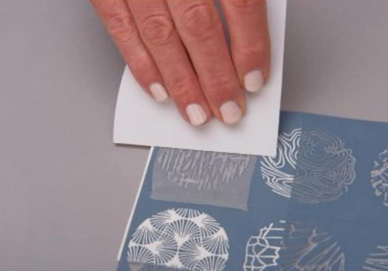 May include: A hand is pressing a white stencil onto a blue surface with white patterned designs. The stencil has a geometric pattern.