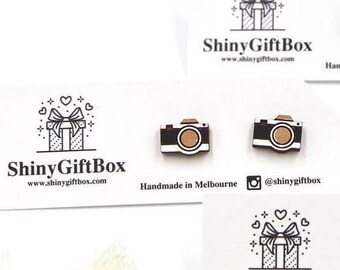 Op de afbeelding: Een paar houten camera-oorbellen met een zwart en bruin ontwerp. De oorbellen zijn handgemaakt in Melbourne, Australië. De tekst "ShinyGiftBox" en het websiteadres "www.shinygiftbox.com" staan ​​gedrukt op de kaart. De social media-handle "@shinygiftbox" is ook inbegrepen.