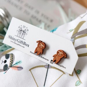 May include: A pair of wooden sloth earrings, handmade in Melbourne, Australia. The earrings are brown and feature a smiling sloth with a bow on its head. The earrings are displayed on a white card with the text "Shiny GiftBox" and a website address.