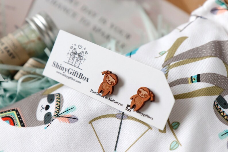 May include: A pair of wooden sloth earrings, handmade in Melbourne, Australia. The earrings are brown and feature a smiling sloth with a bow on its head. The earrings are displayed on a white card with the text "Shiny GiftBox" and a website address.