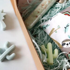 May include: A gift box filled with green and white shredded paper. The box contains a small green cactus, a palm tree, and a white fabric with a sloth design. The fabric has a brown sloth with a colorful headdress and feathers.
