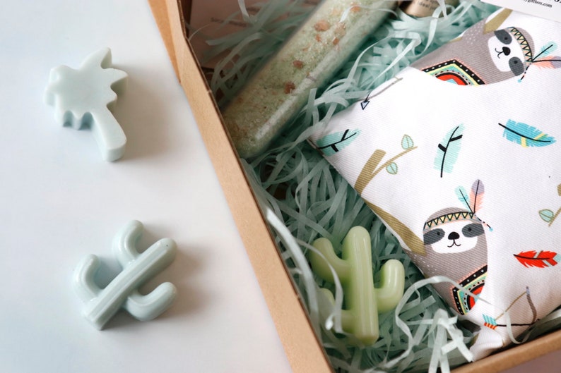 May include: A gift box filled with green and white shredded paper. The box contains a small green cactus, a palm tree, and a white fabric with a sloth design. The fabric has a brown sloth with a colorful headdress and feathers.