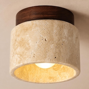 May include: A cylindrical flush mount ceiling light fixture featuring a porous, beige travertine stone shade and a dark brown wooden circular base. The light emits a warm glow from the centre of the stone cylinder, highlighting the natural texture.