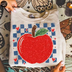 Peut inclure: Un sweat-shirt à col rond beige orné d'un grand motif de pomme rouge à l'aspect vieilli, avec une tige brune et une feuille verte. La pomme est centrée sur un motif à carreaux bleu et beige, agrémenté de petits cœurs rouges et d'étoiles.
