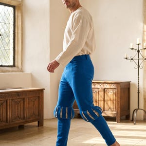 May include: A long-sleeved cream-coloured shirt and bright blue trousers. The trousers feature a fitted design with decorative vertical slits at the knees, revealing a lighter fabric underneath, and a smooth, solid-coloured finish throughout.