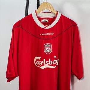 May include: A red short-sleeved soccer jersey featuring a white collar, the Reebok logo in white on the chest, a Liverpool Football Club crest, and the Carlsberg brand name printed in large white letters across the center of the front.