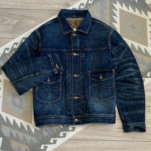 May include: A dark blue denim jacket featuring a classic collar, metal button closures, and two front chest pockets with buttoned flaps. The pockets are detailed with decorative stitching. A brown leather label with the text RRL is visible inside.