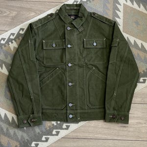 May include: An olive green utility jacket featuring a button-front closure, two chest pockets with button flaps, and two lower slanted pockets. The jacket includes shoulder epaulets, buttoned cuffs, and a collar with a throat latch. RRL label is visible.
