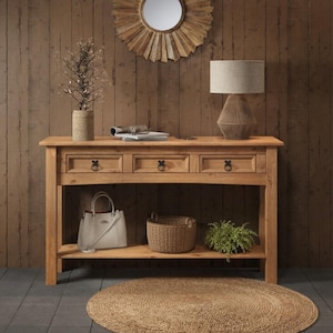 May include: A light brown wooden console table with three drawers featuring dark metal ring pulls. The table has a lower shelf holding a white handbag, a woven basket, and a small green plant. A table lamp with a beige shade sits on top.