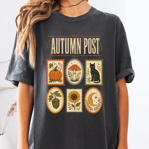 May include: A charcoal gray t-shirt featuring the text AUTUMN POST and six rectangular and oval postage stamp-style illustrations depicting a pumpkin, a mushroom, a black cat, an acorn, a sunflower, and a crescent moon with stars.