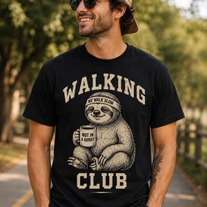 Op de afbeelding: Een zwart t-shirt met korte mouwen en een afbeelding van een luiaard met een hoofdband die een mok vasthoudt. De tekst WALKING CLUB staat boven en onder, met WE WALK SLOW op de hoofdband en NOT IN A HURRY op de mok gedrukt in beige inkt.