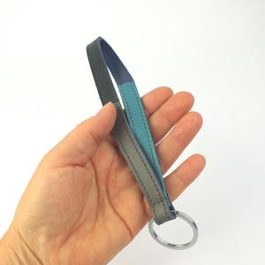 Könnte beinhalten: Ein mehrfarbiger Schlüsselanhänger mit silbernem Schlüsselring. Der Riemen besteht aus Leder in Grau-, Türkis- und Blautönen. Der Schlüsselanhänger wird in einer Hand vor weißem Hintergrund gehalten.