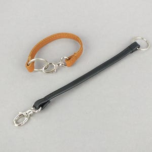 Könnte beinhalten: Ein beiges Lederarmband mit silberfarbenem Karabinerhaken und Spaltring sowie ein längeres schwarzes Lederband mit einem silberfarbenen Karabinerhaken an einem Ende und einem passenden Spaltring am anderen Ende zur sicheren Befestigung.