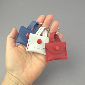 May include: Three miniature leather handbags in red, white, and blue, each with a red button closure and a small handle. The bags are attached to keyrings, ideal for small accessories.