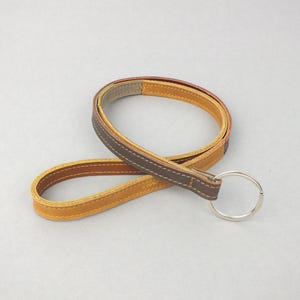 Könnte beinhalten: Ein Schlüsselanhänger aus Leder in Braun- und Beigetönen mit einem silberfarbenen Schlüsselring. Der Schlüsselanhänger ist mit einem Kontrastfaden genäht. Schlichtes und elegantes Design.