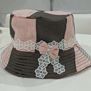 May include: A bucket hat featuring a patchwork design of brown and white houndstooth fabric alternating with solid light pink panels. The hat is decorated with a white floral lace trim encircling the crown and a small pink fabric bow.