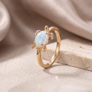 Puede incluir: Un anillo de tono dorado con una piedra ópalo ovalada e iridiscente en el centro, engastada en un marco de metal con forma de tortuga marina. El diseño de la tortuga incluye cuatro patas pequeñas y una cabeza que sobresalen del aro pulido.