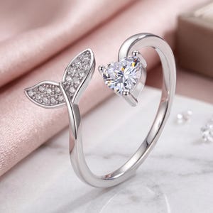 Può includere: Un anello aperto color argento con una pietra preziosa trasparente a forma di cuore su un'estremità e un motivo a coda di pesce incastonato con piccole pietre trasparenti sull'altra. La fascia è lucida e liscia con un'apertura regolabile.