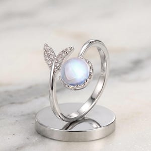Può includere: Un anello aperto color argento con una pietra rotonda bianca iridescente incastonata in una montatura smerlata su un'estremità e un dettaglio a forma di pinna triangolare tempestato di piccole pietre chiare e scintillanti.
