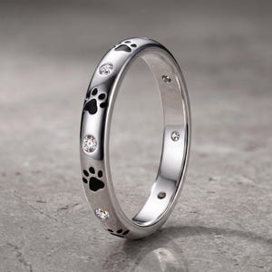 May include: A polished silver-tone ring featuring a repeating pattern of black enamel paw prints alternating with small, clear, round-cut stones set into the band. The metal surface is reflective and smooth, showcasing a continuous circular design.