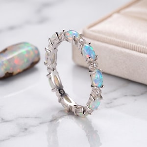 Könnte beinhalten: Ein silberfarbener Ring mit einer Reihe ovaler, schillernder Opale, die sich mit kleinen, klaren, rund geschliffenen Steinen in einer Krappenfassung abwechseln. Das Band zeigt ein durchgehendes Muster dieser Edelsteine um den gesamten Umfang.