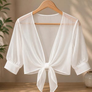 May include: A white sheer cropped bolero with a deep V-neckline, three-quarter length sleeves with cuffs, and a front tie closure. The garment is made of a lightweight, translucent fabric and is displayed on a wooden hanger.