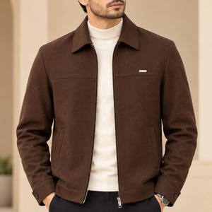 May include: A dark brown zip-up jacket with a classic collar, long sleeves, and a small metallic rectangular detail on the chest. The jacket is worn over a cream-colored mock neck sweater and paired with black trousers.