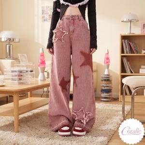 May include: Wide-leg denim jeans in a faded dusty rose color featuring a high-waisted fit, a button and zipper closure, and large star-shaped patches with frayed edges on the front, paired with red and white sneakers.
