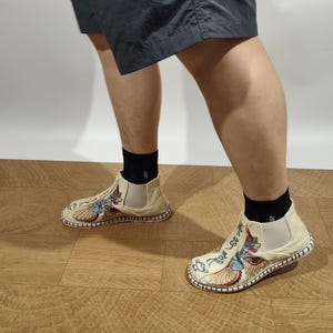 May include: Off-white slip-on shoes featuring intricate embroidery of a stylized creature and Asian-inspired calligraphy on the upper. The shoes have elastic side panels, a rounded toe, and a stitched rope-textured trim along the outer sole edge.