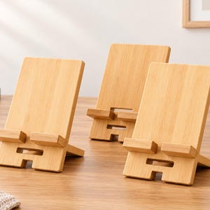 May include: Three light brown bamboo phone stands with a rectangular back panel and a horizontal ledge for device support. Each stand features a cutout slot at the base and a hinged support arm on the back to maintain an upright angled position.
