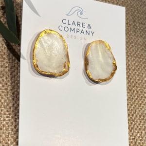 May include: A pair of stud earrings featuring irregular, white, translucent, stone-like shapes with textured, metallic gold-painted edges. The earrings are attached to a white display card printed with the text CLARE & COMPANY DESIGN.
