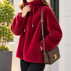 Puede incluir: Una chaqueta de color rojo intenso, manga larga y textura afelpada, con cuello alto, cremallera frontal y cordones ajustables a juego con puntas metálicas. Se combina con un bolso de hombro de cuero marrón oscuro con un emblema metálico dorado.