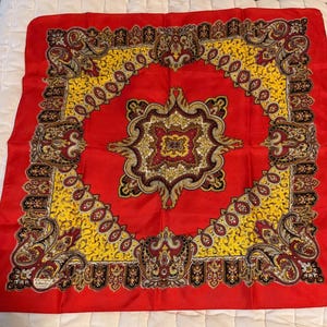 May include: A square scarf featuring a vibrant red background with a central ornate medallion pattern in yellow, black, and white. The border displays intricate paisley and floral motifs in matching colors, with a small label in the corner.