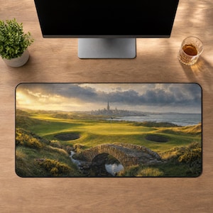 May include: A rectangular desk mat featuring a landscape print of a golf course with a stone bridge in the foreground, a green fairway, sand bunkers, and a distant coastal town under a cloudy, golden sunset sky.
